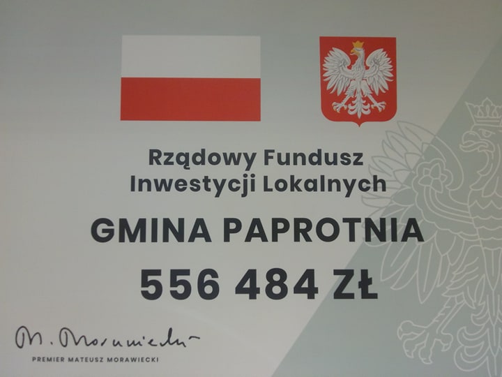 Obraz przedstawia tablice informacyjną. Na gorze tablicy widnieje flaga oraz godło Polski , poniżej napis Rządowy Fundusz Inwestycji Lokalnych, Gmina Paprotnia oraz kwota 556 484 zł 