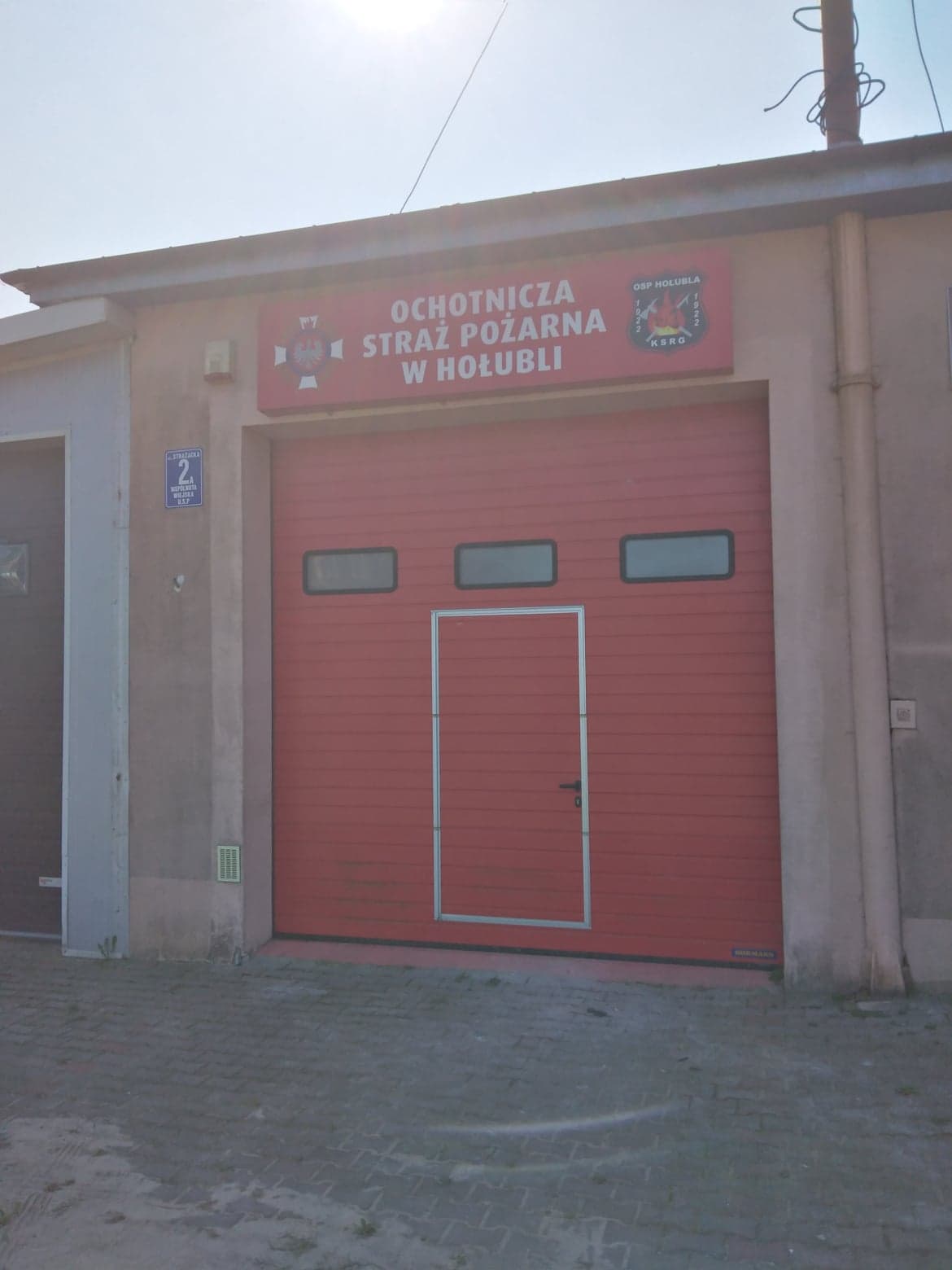 Zdjęcie przedstawia strażnicę OSH Hołubla od zewnątrz. Widoczne są czerwone drzwi oraz duży szyld z napisem Ochotnicza Straż Pożarna w Hołubli
